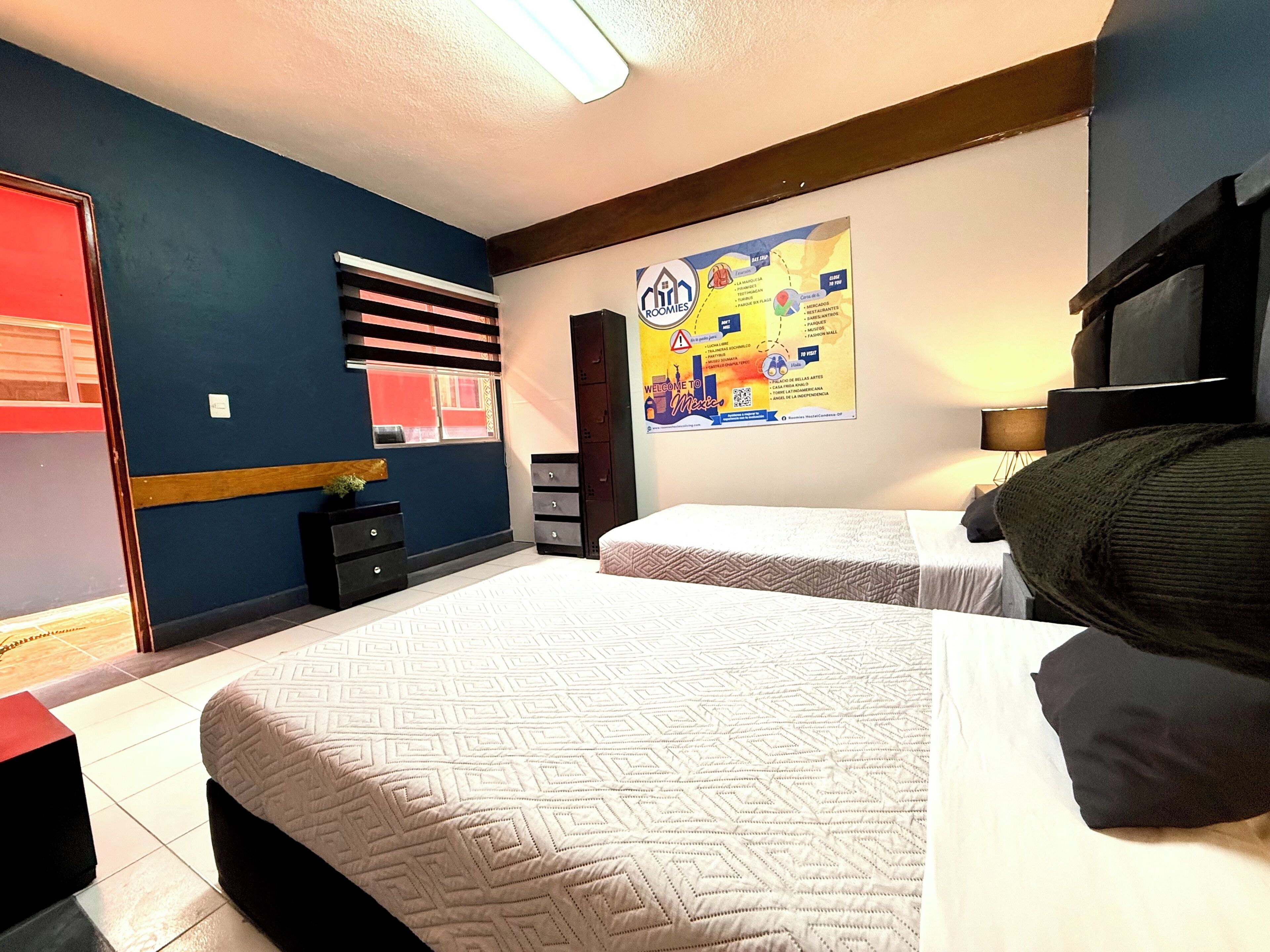 Une chambre d’hôtel avec deux lits, une grande affiche au mur et une fenêtre avec des stores.