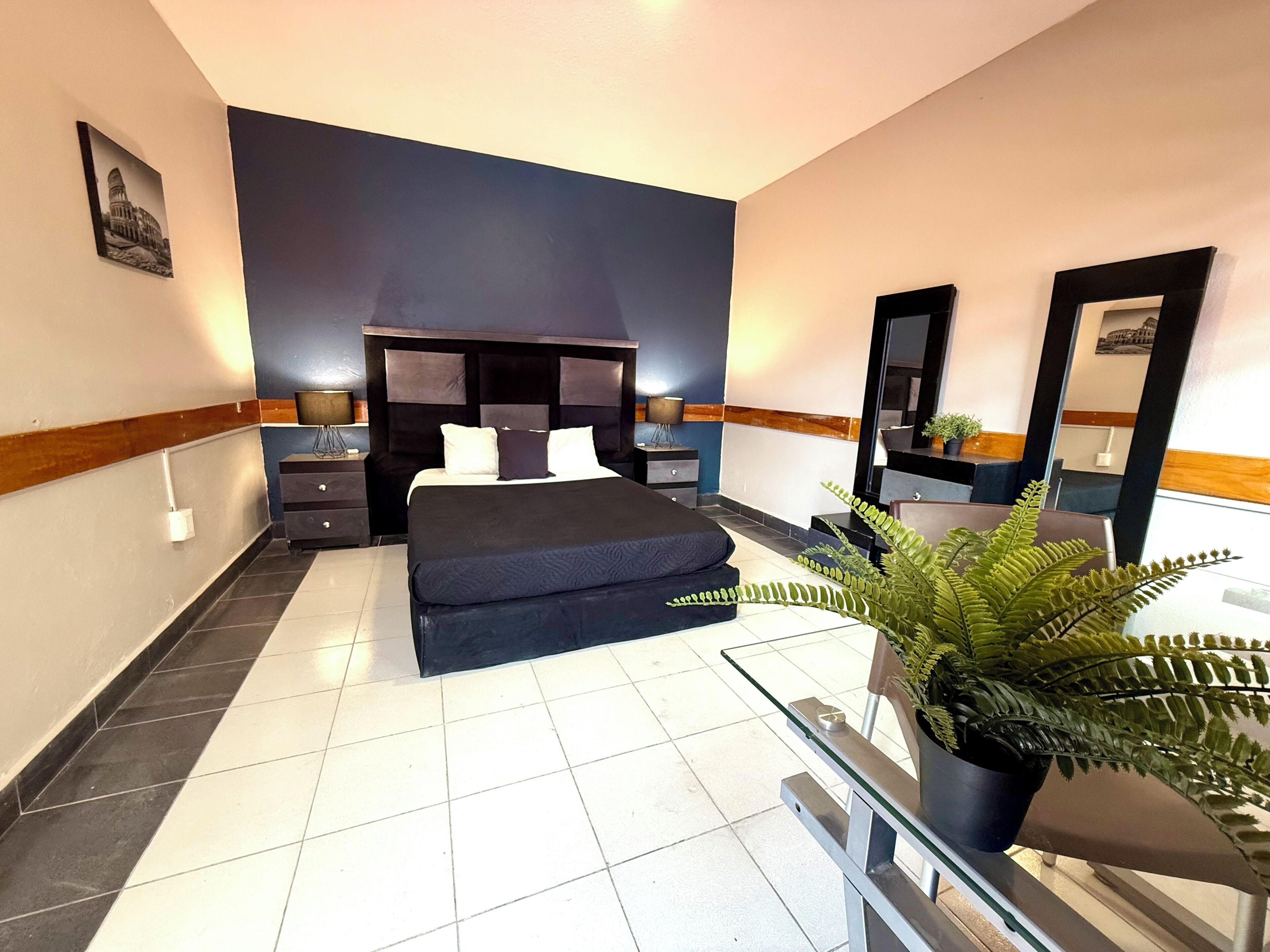 A modern bedroom with a large bed, a nightstand, a dresser, a mirror, and a potted plant.