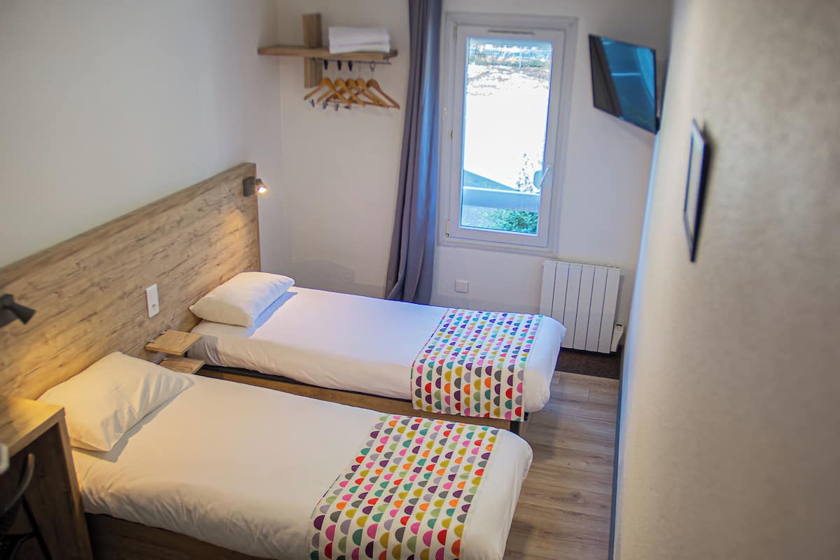 A bedroom with two single beds, a wooden headboard, a bedside table, a window with curtains, and a mounted TV.