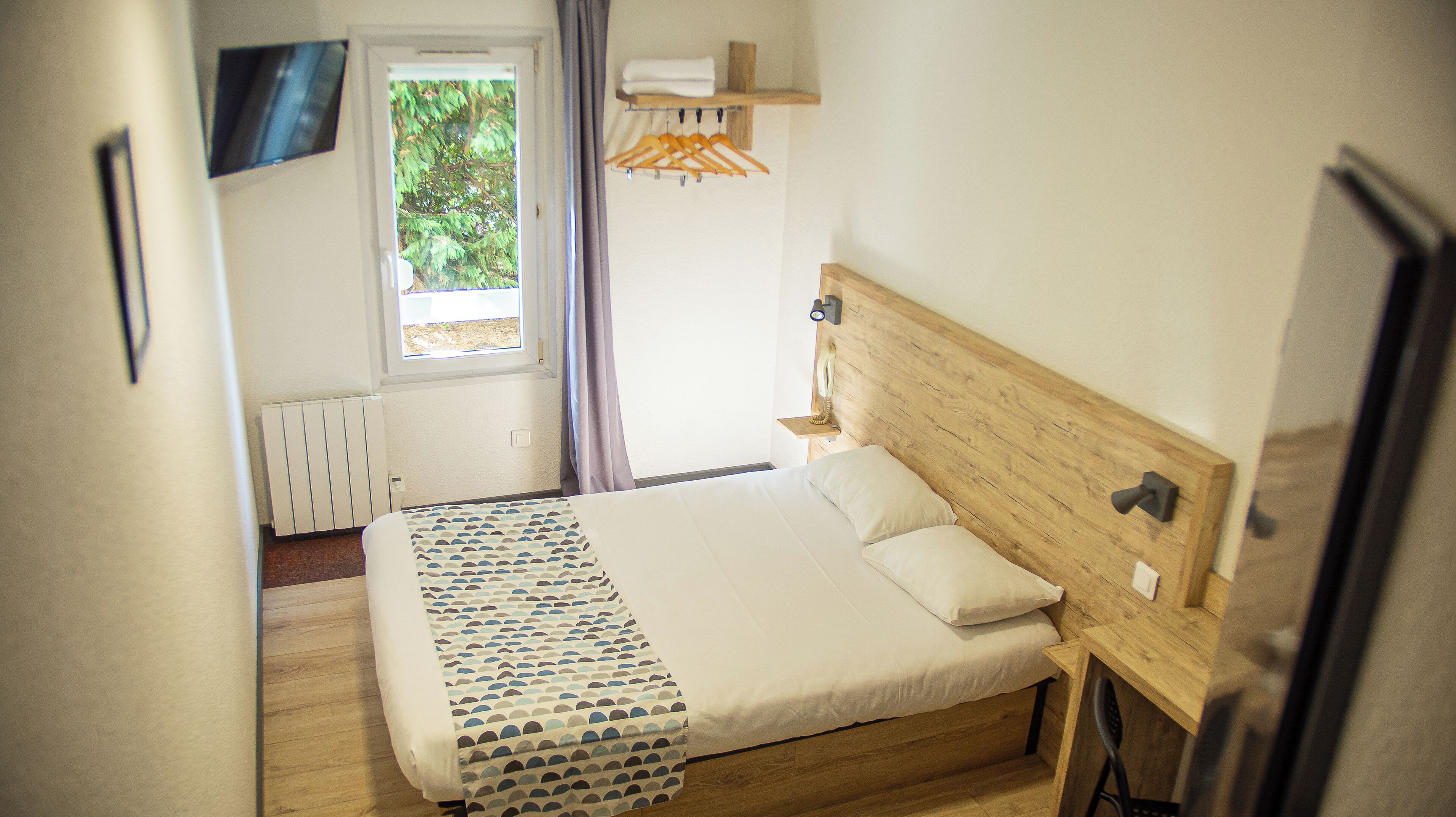 A neatly made bed with a patterned bedspread, a bedside table with a lamp, and a window with a view of trees.