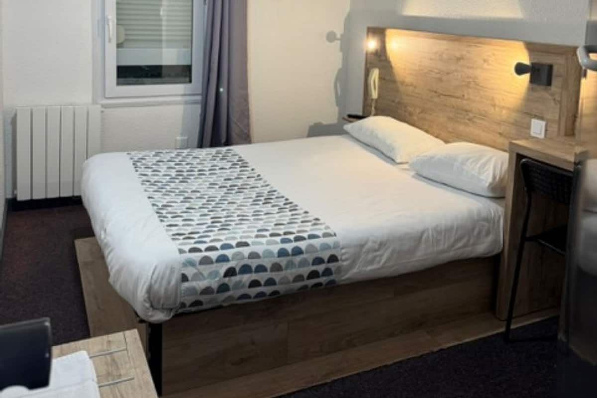 A hotel room with a bed, a desk, a chair, a TV mounted on the wall, and a window with curtains.
