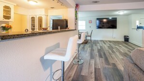 A modern kitchen with a granite countertop, white bar stools, and a dining area with a TV and wooden flooring. - 8 Min ATandT Stadium | Central 4BR Retreat (Grand Prairie)