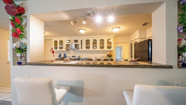 A modern kitchen with white cabinets, a granite countertop, and a stainless steel refrigerator. There are two white chairs in front of the kitchen. - 8 Min ATandT Stadium | Central 4BR Retreat (Grand Prairie)