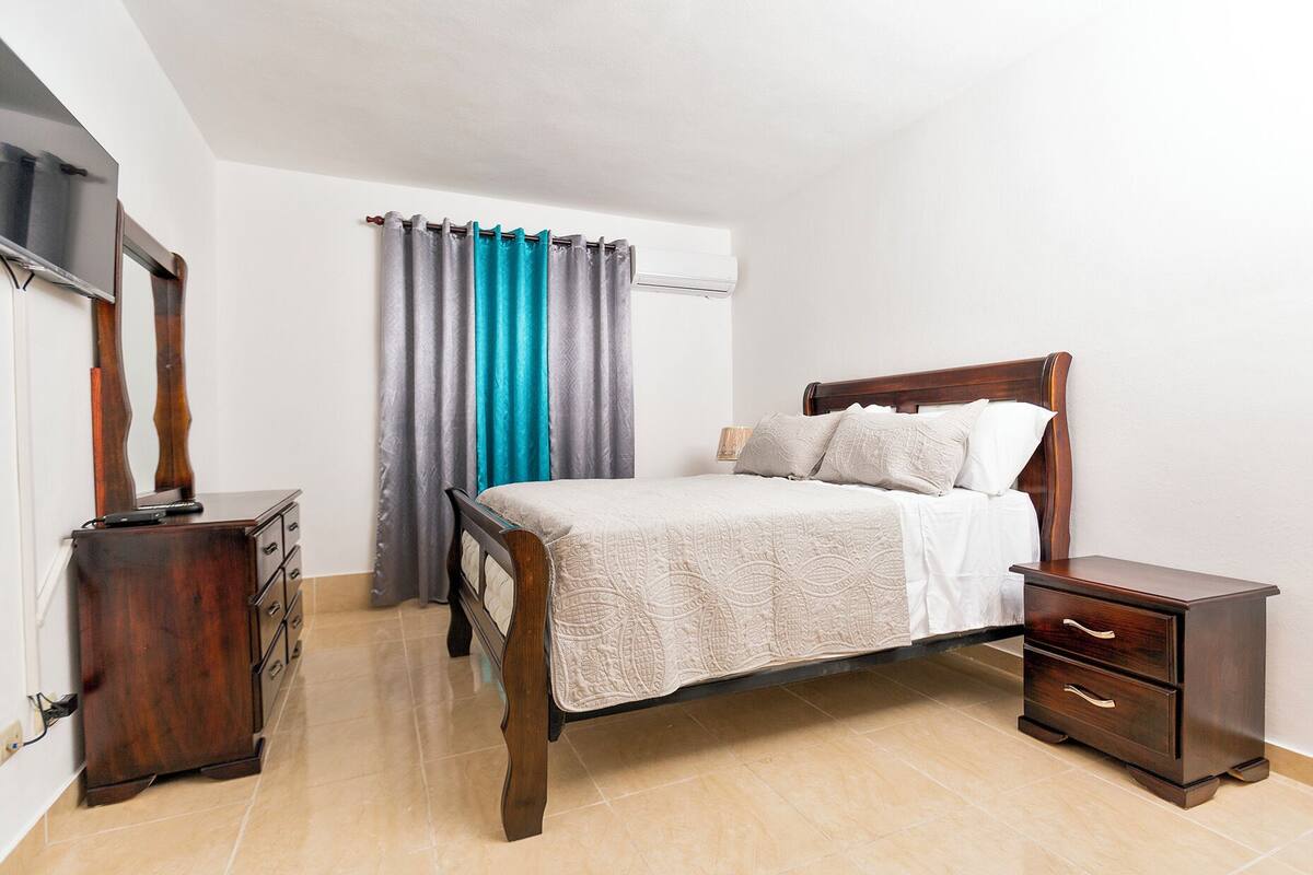 A bedroom with a wooden bed, a nightstand, a dresser, and a window with curtains.