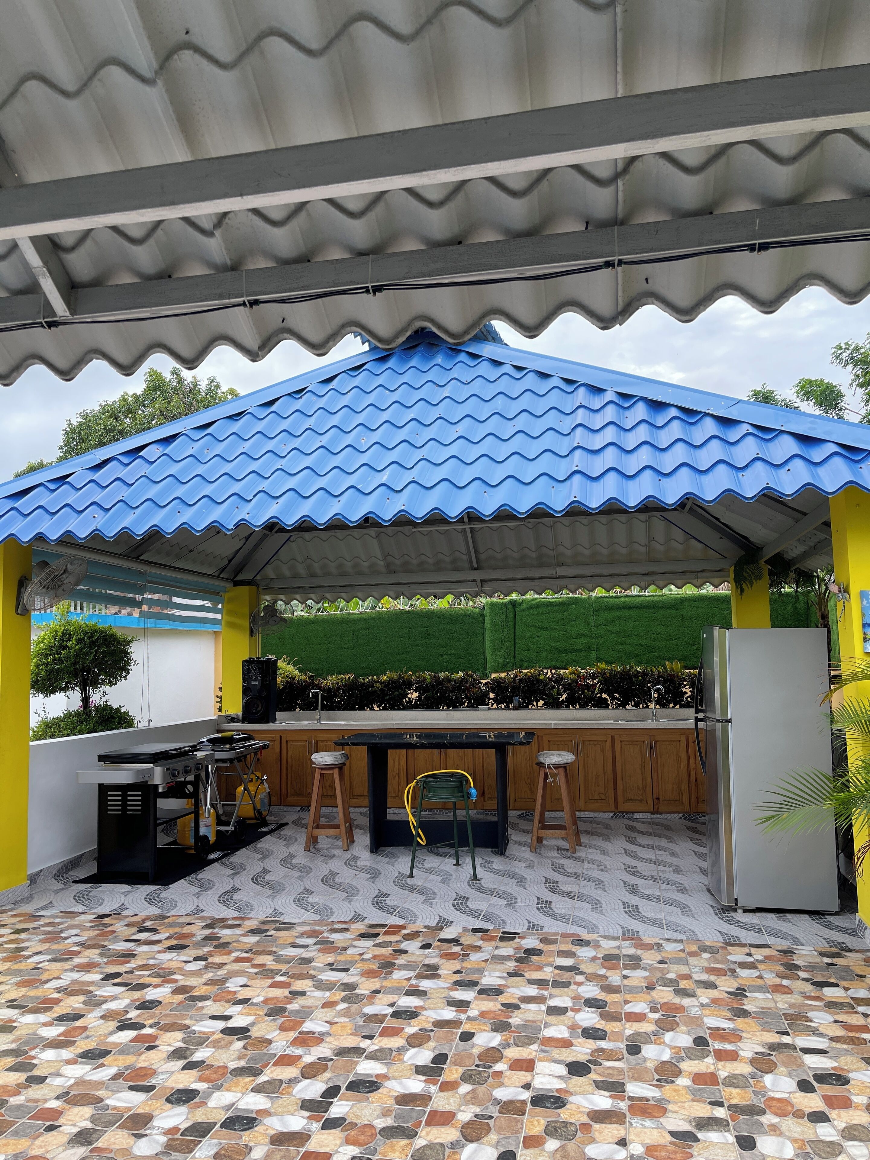 An outdoor kitchen area with a blue corrugated roof, a tiled floor, and a green artificial grass backdrop.