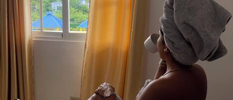 A person sitting on a bed with a towel wrapped around their head, a window with curtains, and a view of greenery outside.