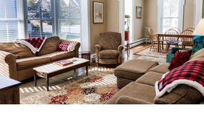 A living room with a sofa, armchair, and ottoman, a dining table with chairs, and large windows with blinds. - Cozy Ludlow, Vermont Farmhouse Ski Retreat with hot tub and fire pit (Ludlow)