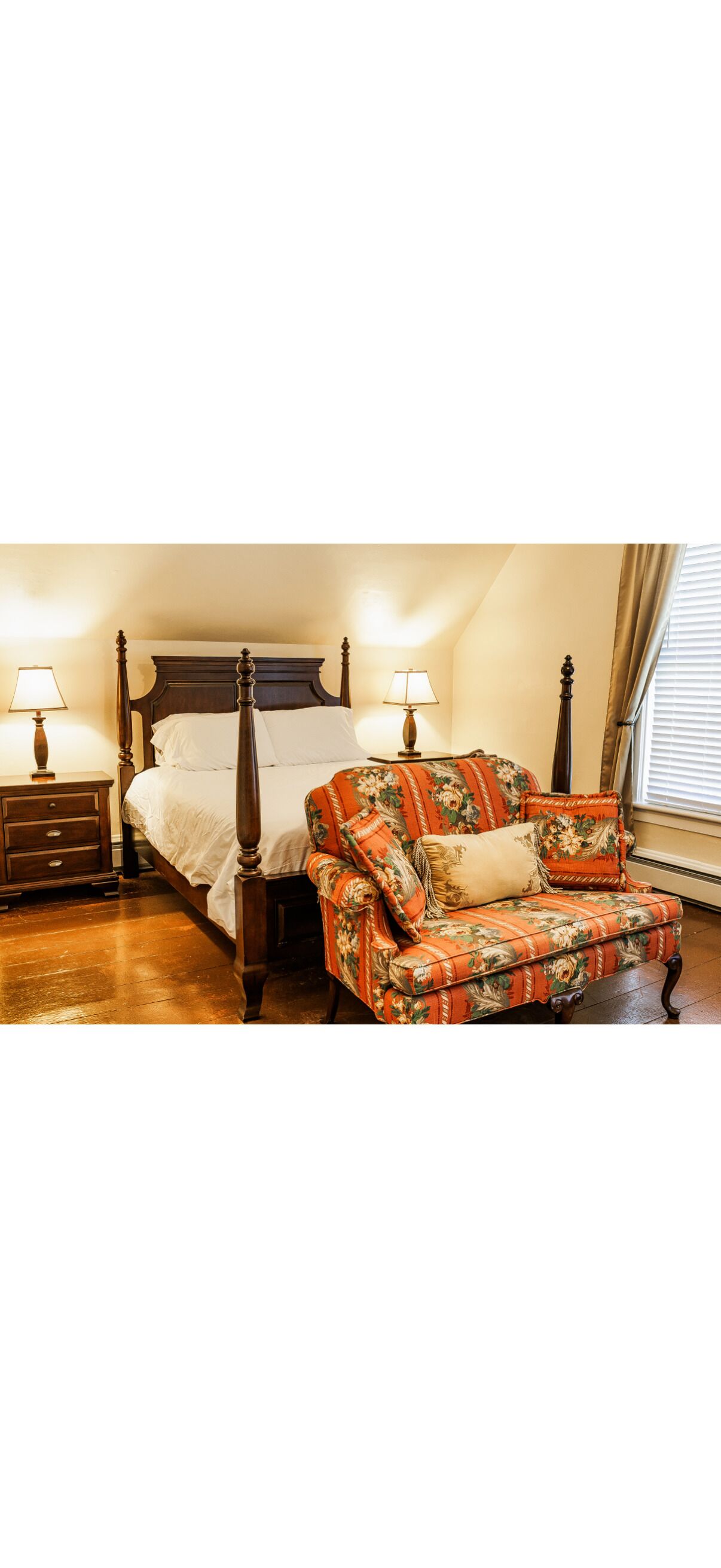 A four-poster bed with white linens, a wooden nightstand with a lamp, and a floral-patterned armchair with a cushion.