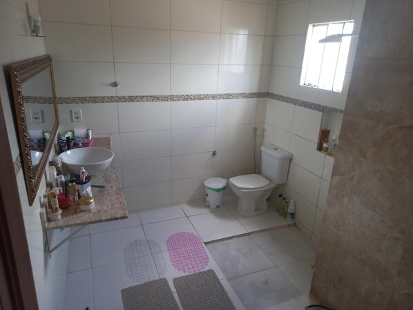 Une salle de bain avec un lavabo, des toilettes et un miroir, tous de couleur blanche. On trouve des articles de toilette sur le comptoir et une fenêtre qui laisse entrer la lumière naturelle.