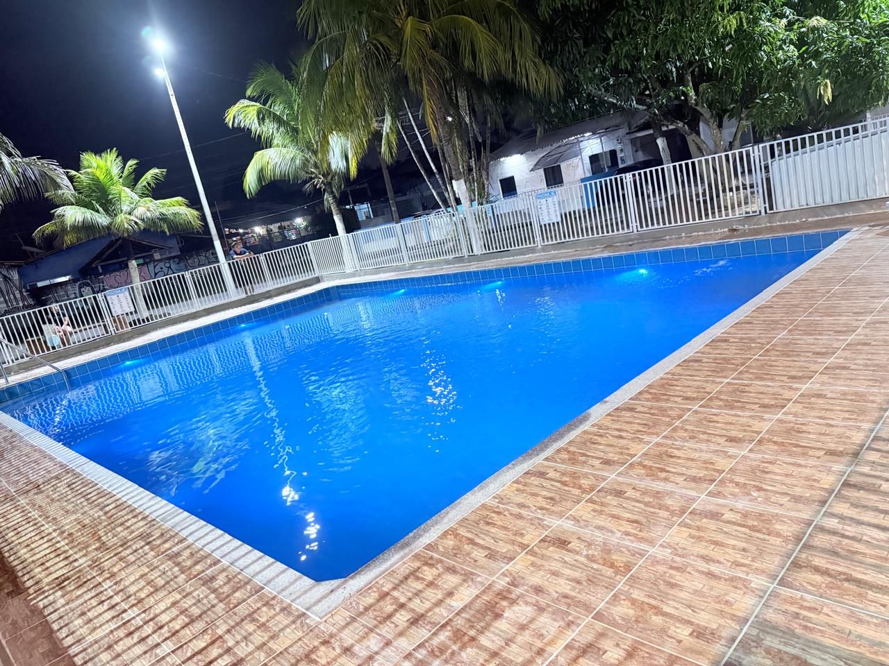 Una piscina rectangular con borde de azulejos, rodeada por una valla blanca y palmeras.