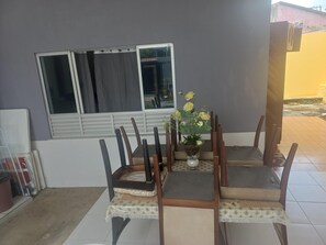 Un coin repas avec une table dressée pour quatre personnes, une plante en pot aux fleurs jaunes et une fenêtre avec des rideaux.