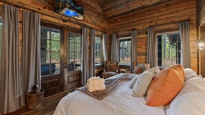 A wooden cabin room with a bed, curtains, a television, and a window with a view of trees. - Hot tub Sauna King • Fireplace • Pet‑Friendly (Broken Bow)