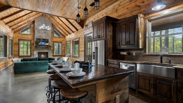 A spacious kitchen with wooden beams, a stone fireplace, and a large island with bar stools. - Hot tub Sauna King • Fireplace • Pet‑Friendly (Broken Bow)
