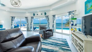Een moderne woonkamer met een groot raam dat uitzicht biedt op de oceaan, een zwarte leren bank, een flatscreen-tv en een plafondventilator.