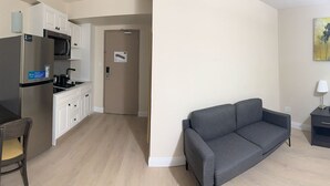 Un espacio habitable compacto con kitchenette, una mesa de comedor con una silla, un sofá gris, una mesita auxiliar y un cuadro en la pared.