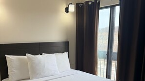 Una cama bien hecha con ropa blanca y un edredón beige estampado, cabecera oscura y una ventana con cortinas.