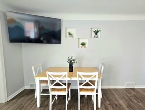 Ein Essbereich mit einem Holztisch und weißen Stühlen, einem an der Wand befestigten Flachbildfernseher und kleinen, in Töpfen stehenden Pflanzen an der Wand.