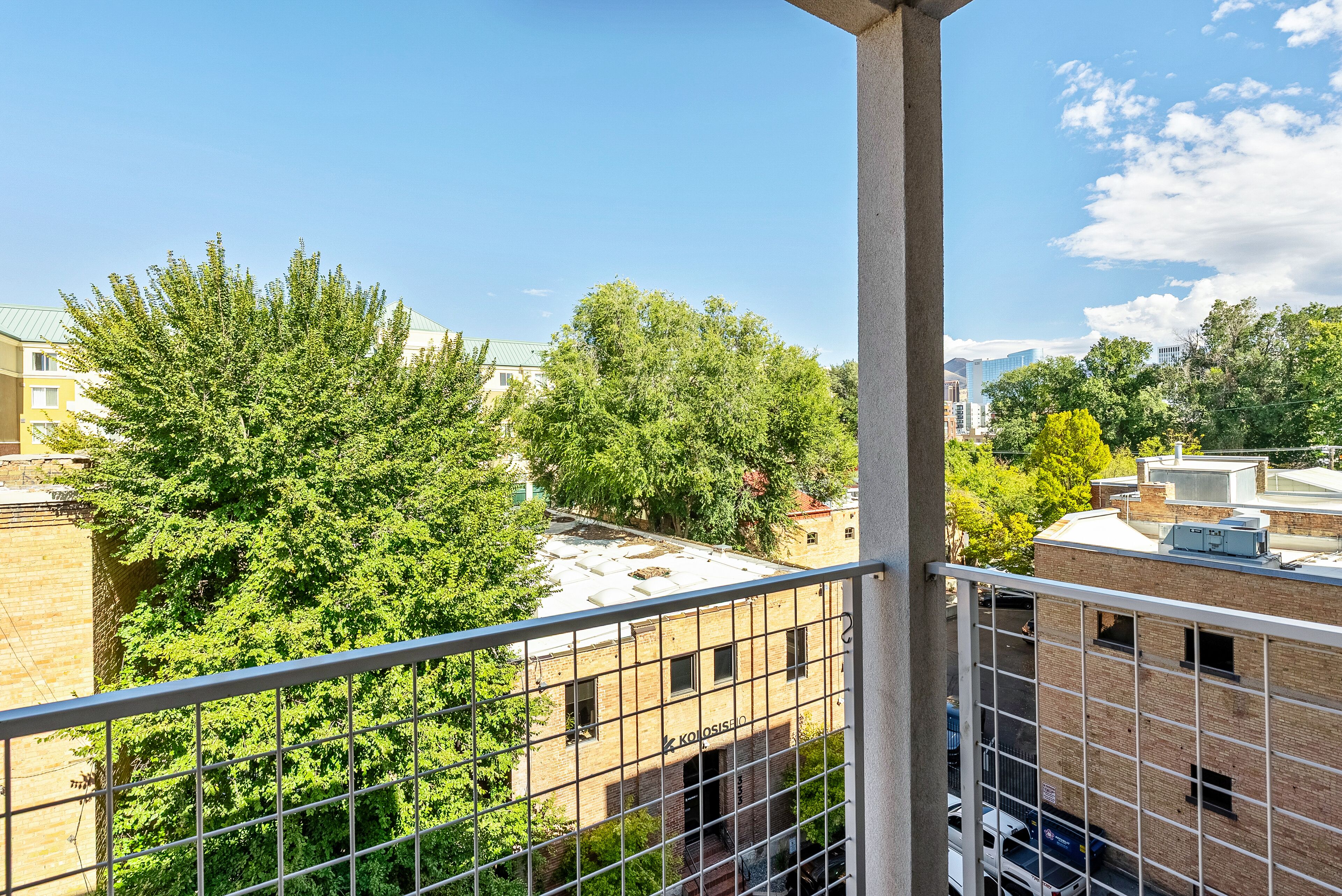 A view from a balcony overlooking a brick building with a visible sign, surrounded by trees and other buildings.