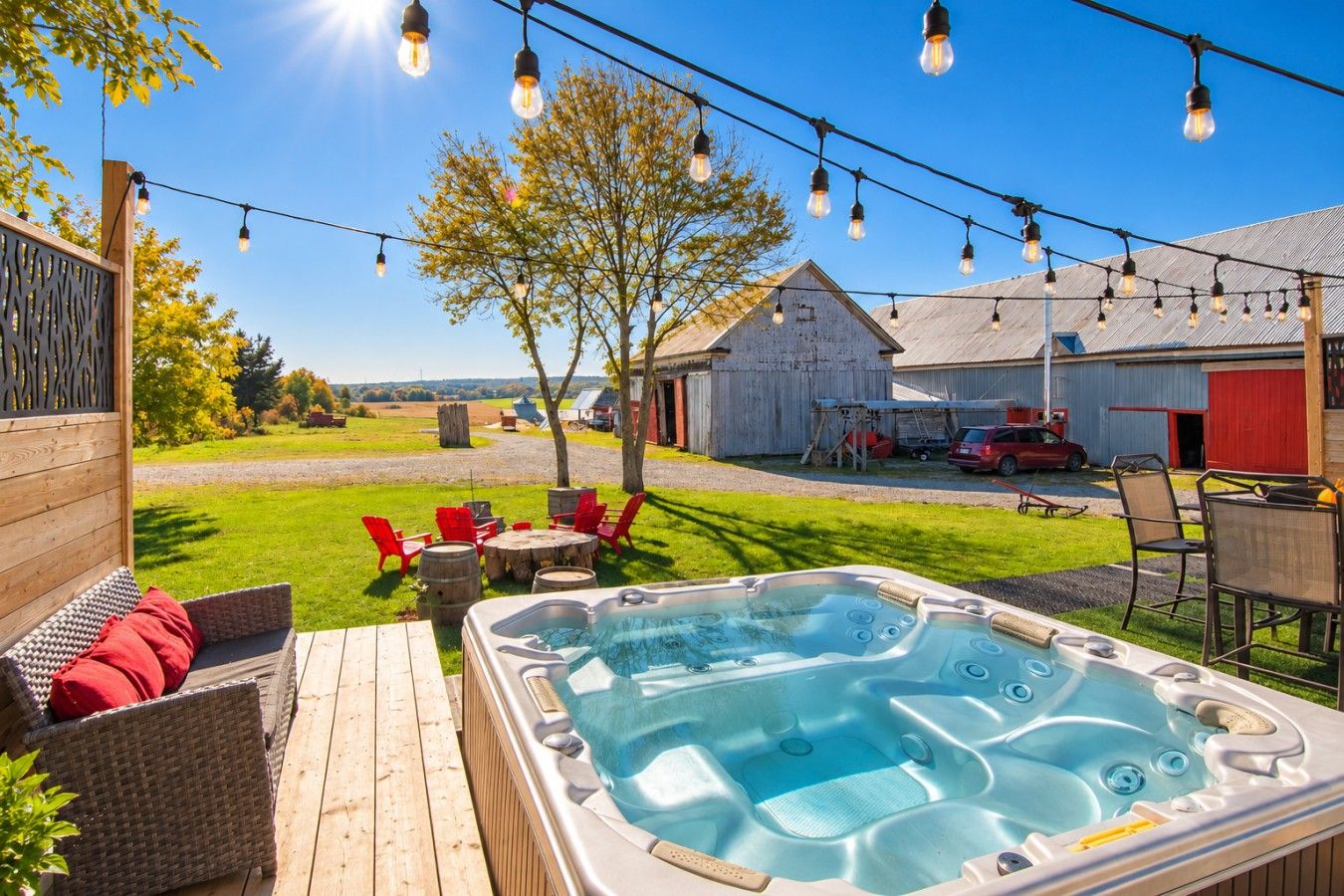 A backyard with a hot tub, outdoor seating, string lights, and a barn.