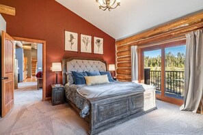 Ein Schlafzimmer mit einem großen Bett, einem hölzernen Kopfteil, einer Kristallleuchtre und Blick auf einen Wald durch große Fenster.