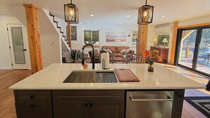 Cocina con isla central, armarios de madera y lavavajillas de acero inoxidable. Zona de comedor con sofá y mesa, y una escalera que lleva a otro nivel.