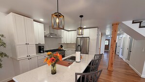 Una cocina moderna con armarios blancos, un refrigerador de acero inoxidable, una mesa de comedor de madera con sillas y lámparas colgantes.