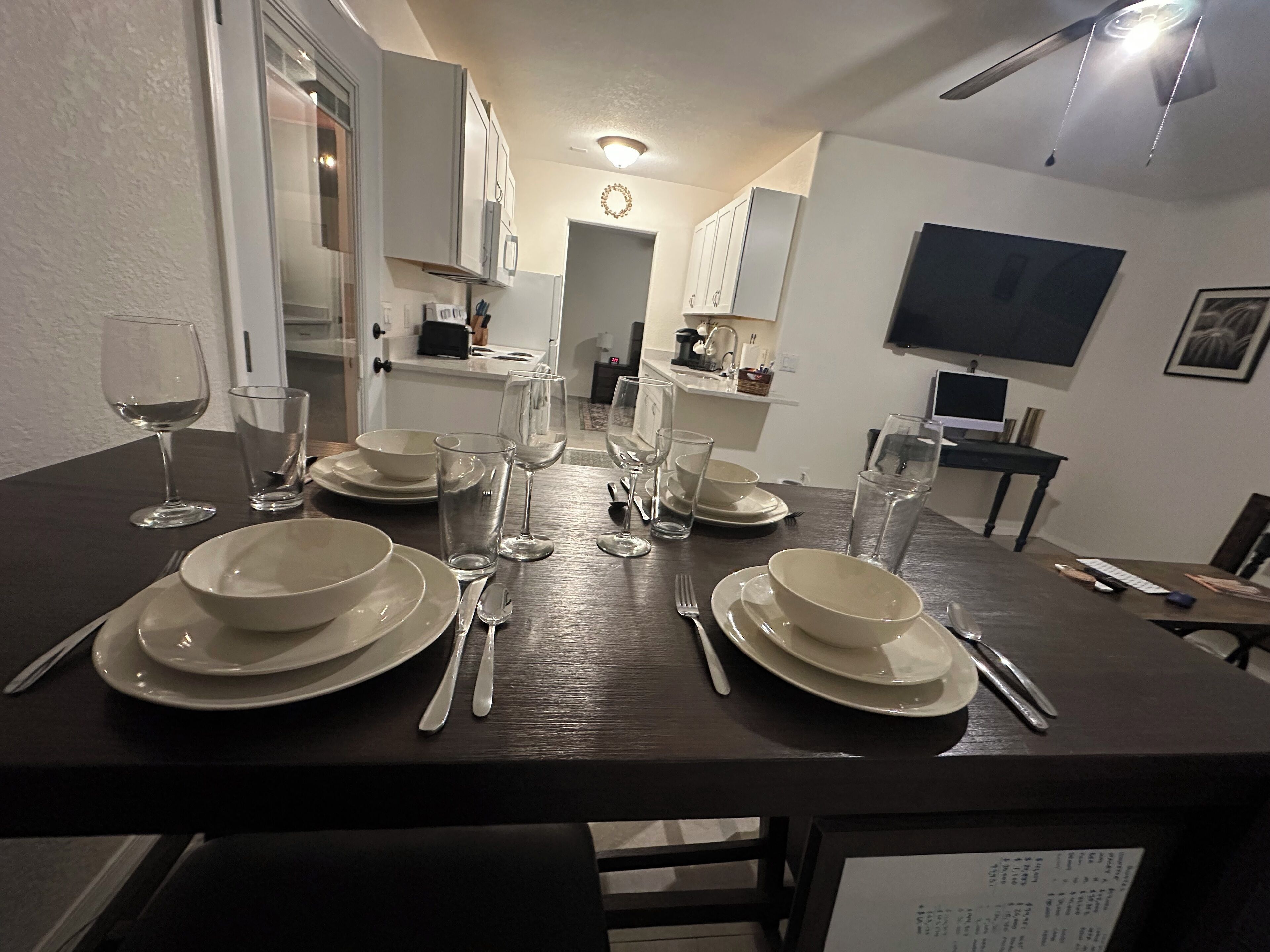 Une table à manger dressée avec des assiettes, des verres et des couverts, avec une cuisine et un téléviseur visibles à l’arrière-plan.
