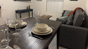 A dining area with a dark wooden table set for two, a flat-screen TV mounted on the wall, a small side table, and a window with blinds. - Southwest-Chic 1BR Casita, Private Yard + Driveway, Northern Phoenix Getaway (Glendale)