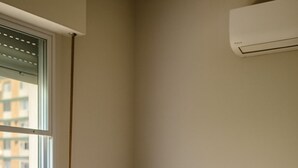 Une petite chambre avec un lit simple, une fenêtre, un climatiseur fixé au mur et un mur uni.