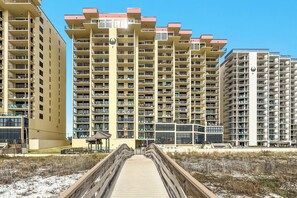 A multi-story residential building with balconies, a wooden boardwalk leading to the entrance, and a clear sky. - Phoenix 2 - 2044 | Beachfront | Outdoor & Indoor Pool | Tennis (Orange Beach)