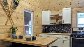 Ein Holzinnenraum mit einer Küchenzeile, die weiße Schränke, eine schwarze Arbeitsplatte und einen Herd zeigt. An der Küchenarbeitsplatte stehen zwei Hocker und über der Spüle befindet sich ein Fenster.