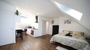 A modern loft bedroom with a sloped ceiling, a built-in kitchen, and a large bed with patterned bedding. - Eden (Cambrai)