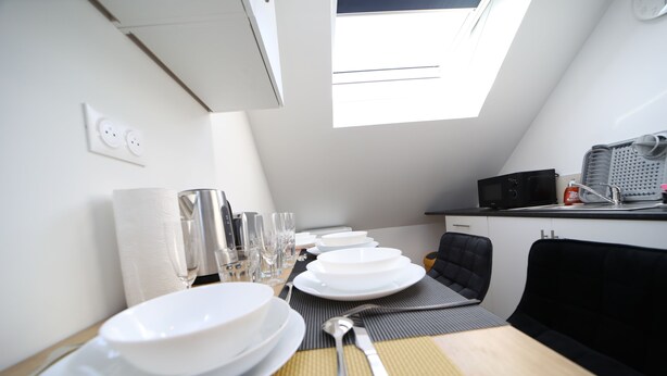 A dining area with a table set for four, including plates, glasses, and cutlery. A black chair is positioned at the table, and a microwave is visible in the background. - Eden (Cambrai)
