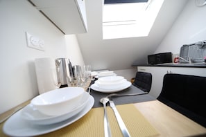 Un coin repas avec une table dressée pour quatre personnes, comprenant des assiettes, des verres et des couverts. Une chaise noire est placée à la table, et un four à micro-ondes est visible à l’arrière-plan.