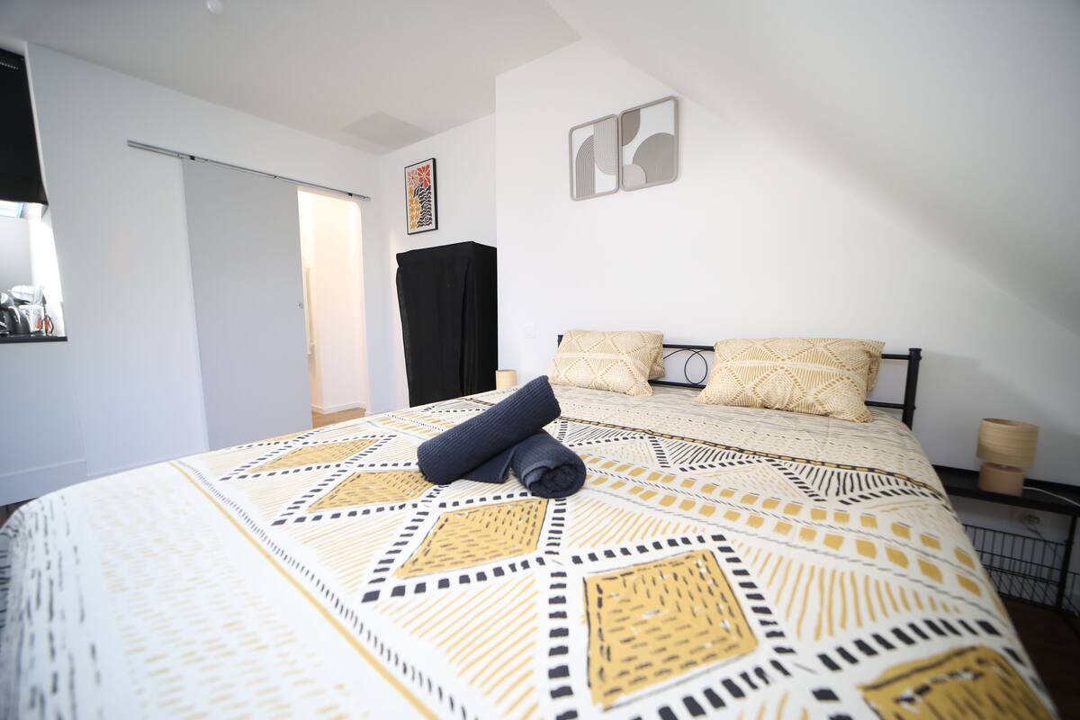 Quarto com colcha estampada, toalha enrolada na cama e um guarda-roupa preto ao fundo.