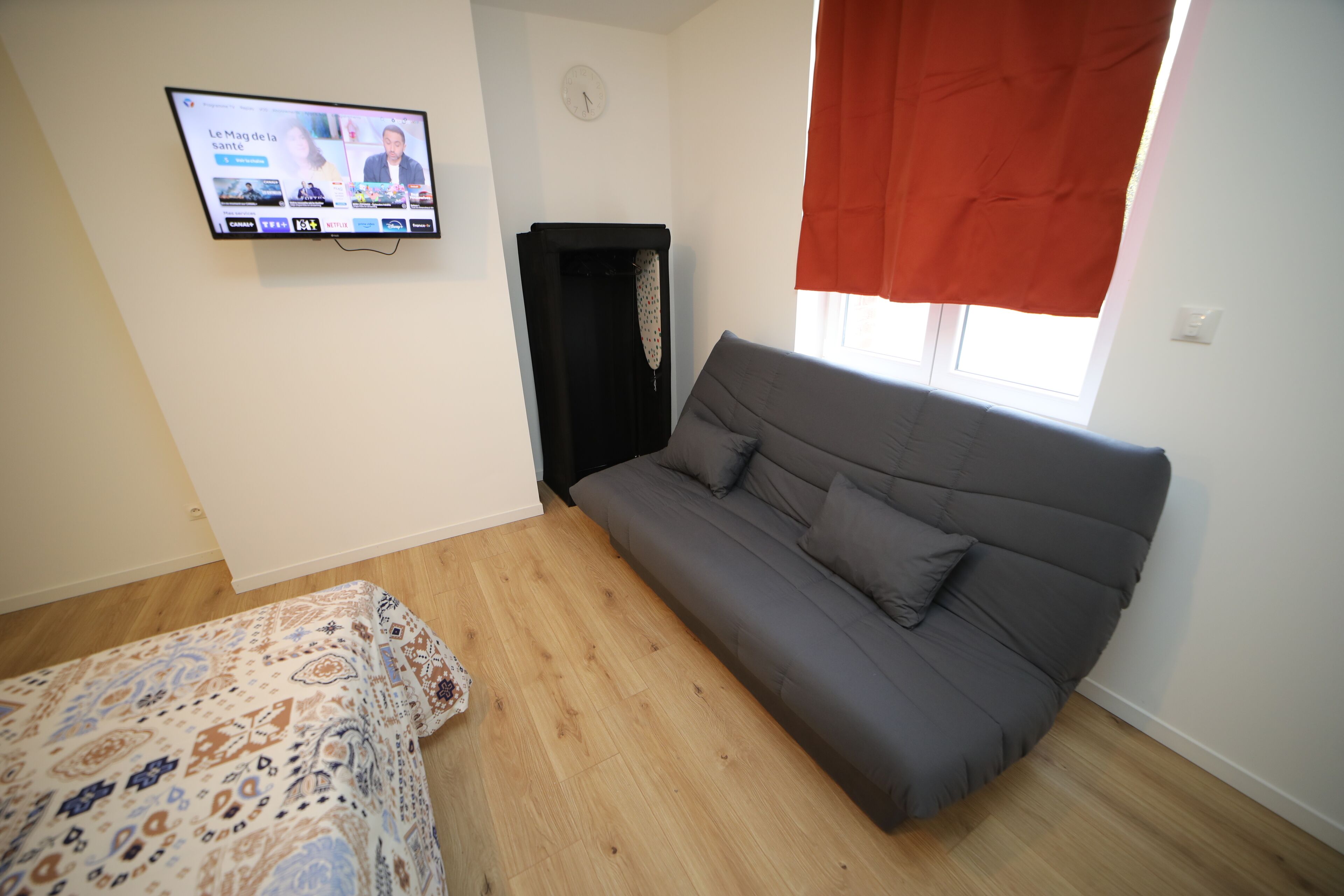 A room with a grey sofa, a patterned bedspread, a television displaying a news program, and a window with a red curtain.