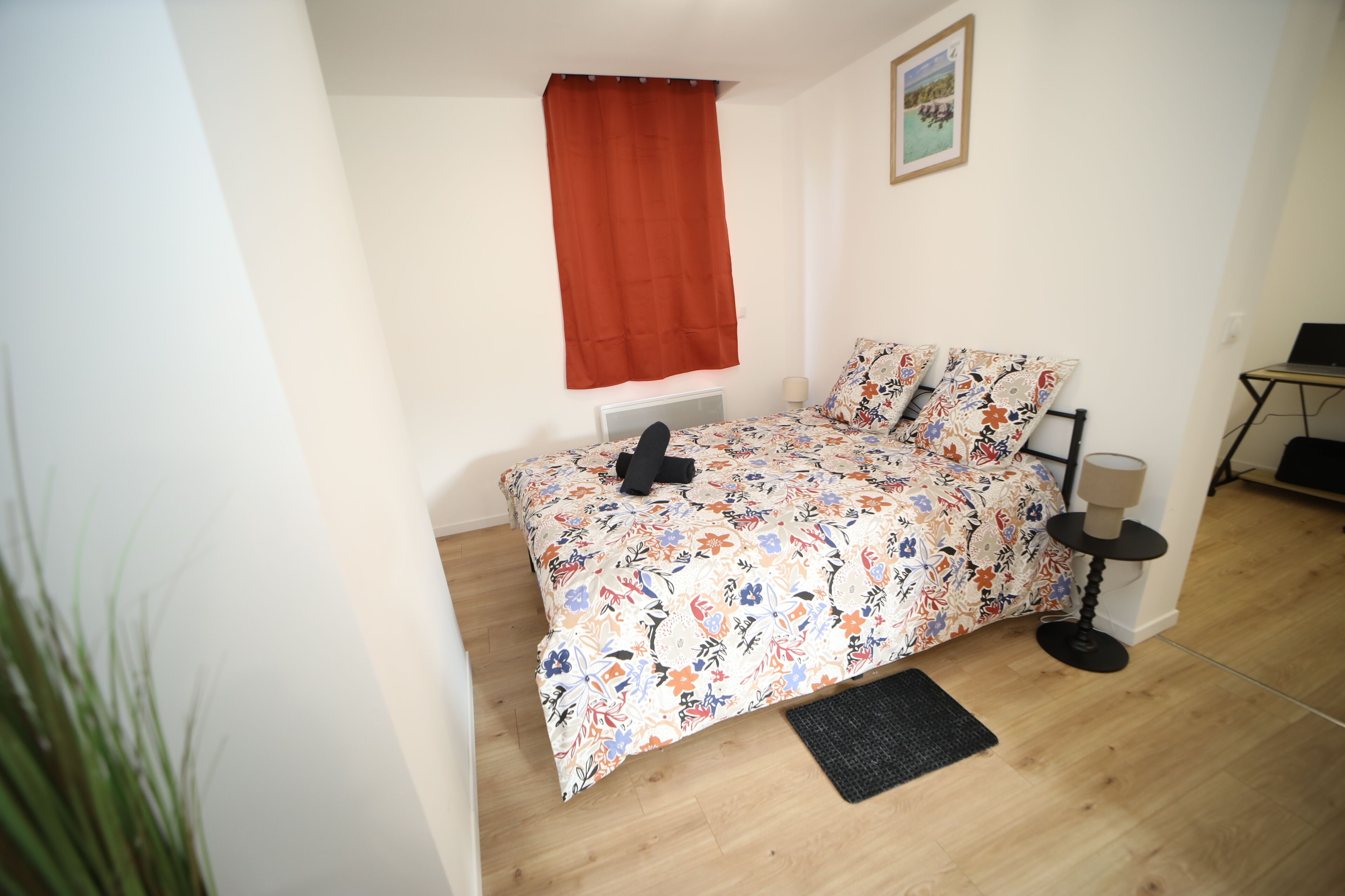 A bedroom with a floral bedspread, a red curtain, a small bedside table with a lamp, and a laptop on a desk.