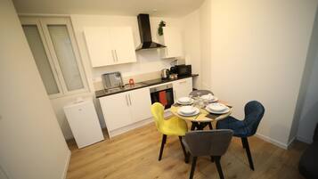 Une cuisine compacte avec des meubles blancs, une hotte noire, un four à micro-ondes et un petit réfrigérateur. On y trouve une table à manger avec des chaises, ainsi que quelques assiettes et tasses.