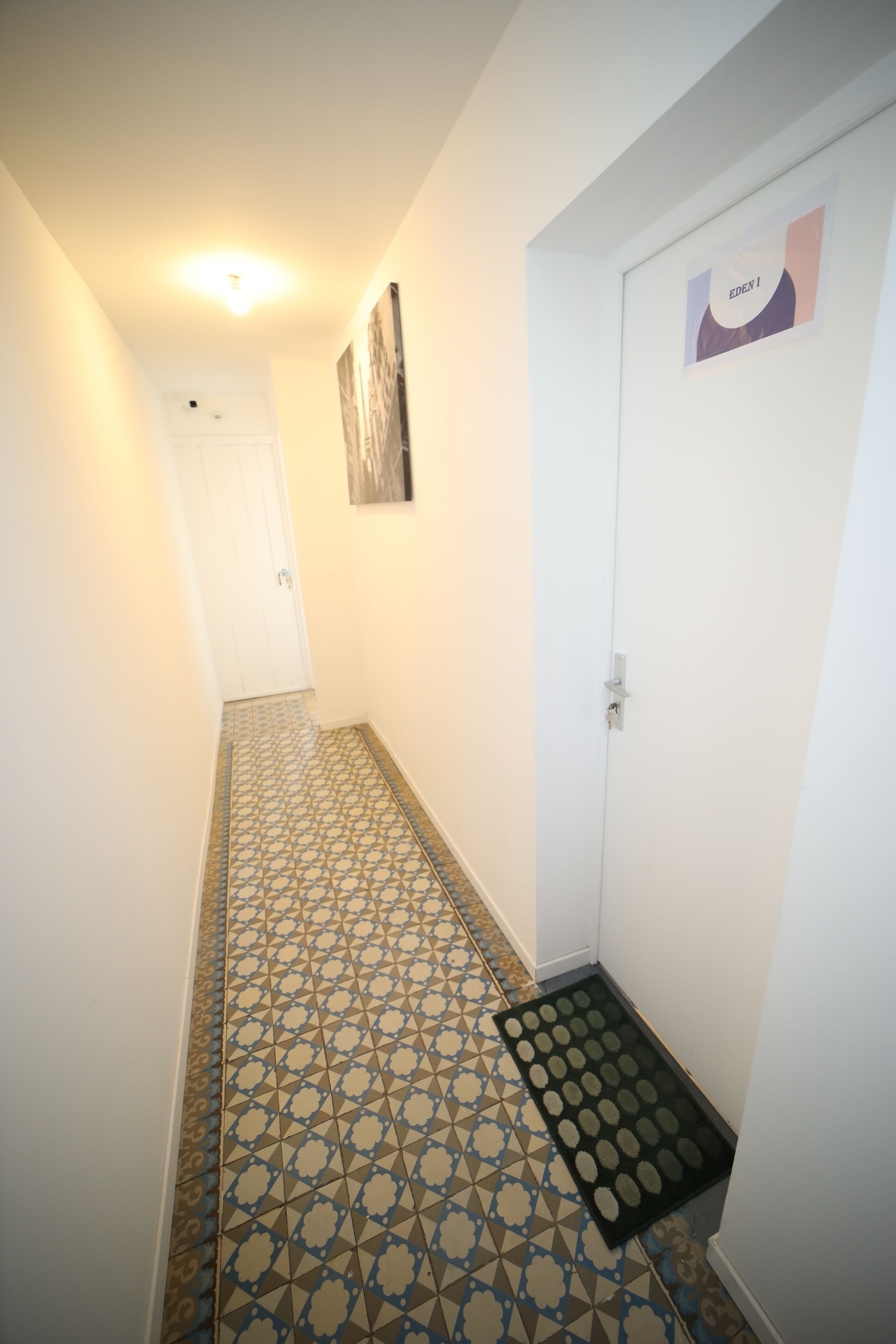 Un couloir étroit avec des carreaux de sol à motifs, une porte portant une plaque avec un nom, et un tableau accroché au mur.