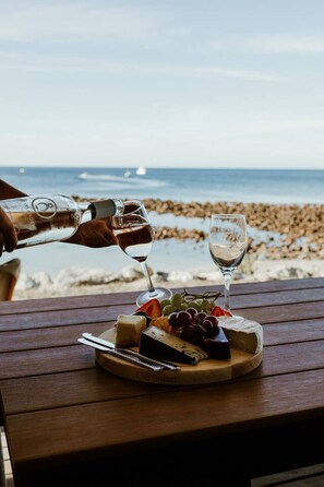 Une table en bois avec une planche de fromages, des raisins et un verre de vin, le tout installé sur une plage, avec l’océan et un voilier en arrière-plan.