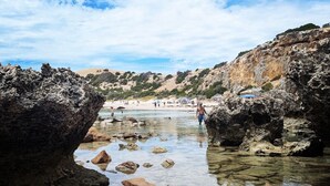 Uma praia rochosa com águas claras e uma vista distante de uma praia de areia.