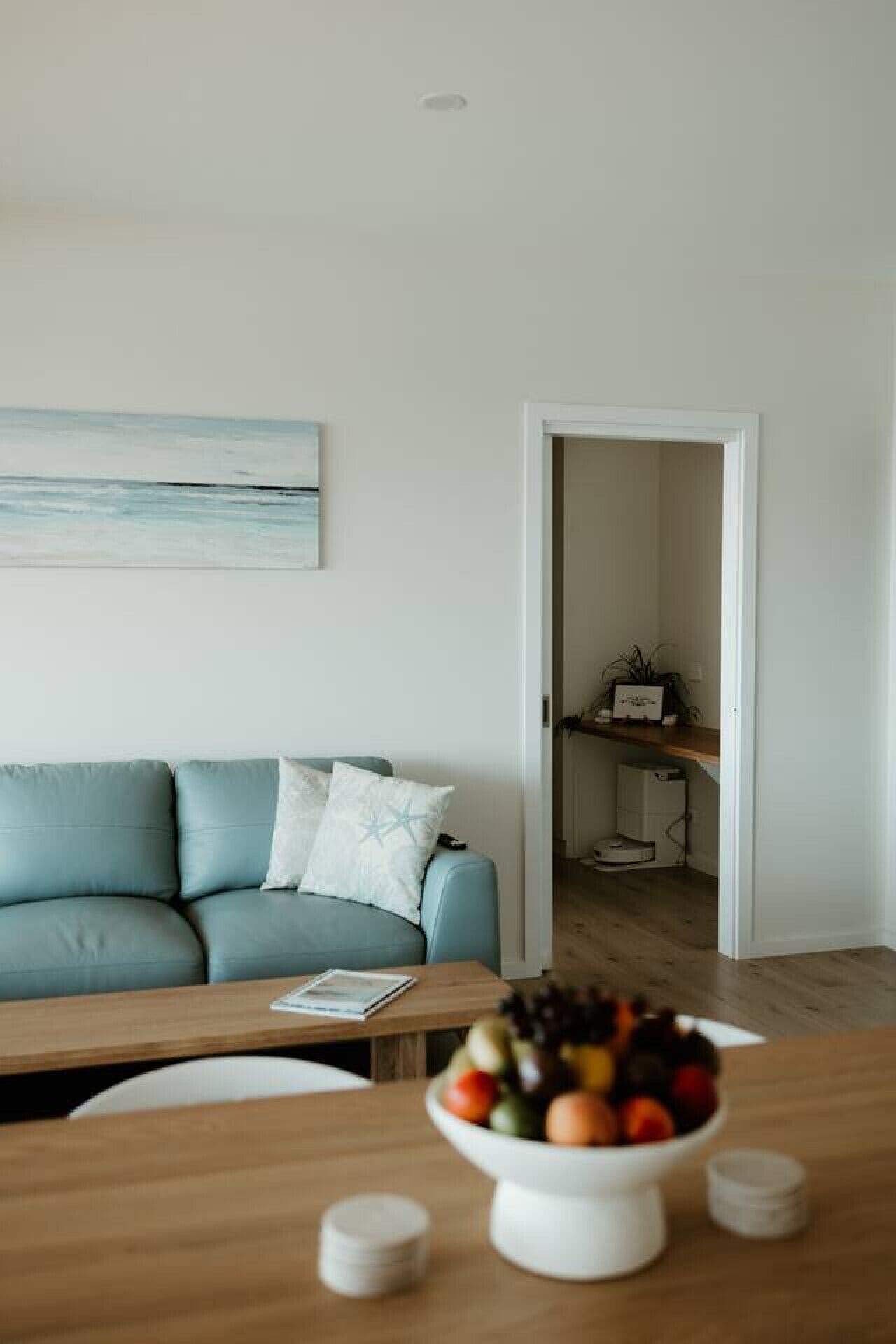 Ein Wohnzimmer mit einem blauen Sofa, einem Couchtisch aus Holz mit Obstschale und einem Bild an der Wand.