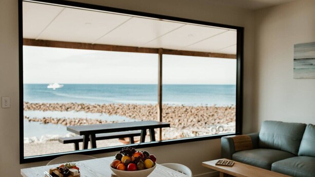 Quarto com uma grande janela com vista para uma praia, uma mesa de madeira com uma cesta de frutas e um sofá cinzento.