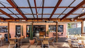 Un café al aire libre con mesas y sillas de madera, una planta de cactus y una estructura de techo metálico.