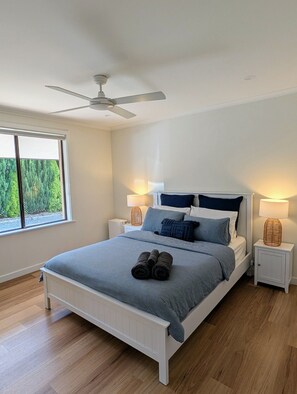 A bedroom with a bed, two bedside tables, a ceiling fan, and a window with a view of greenery. - Sails: Family-friendly, in the heart of town (Kingscote)