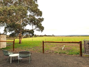 A fenced outdoor area with a table and chairs, a tree, and a view of a green field.
