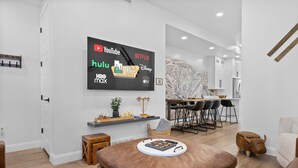 En moderne stue med et fladskærms-tv der viser streamingtjenester, et spiseområde med et bord og stole, og et hyggeligt opholdsområde med en skammel.