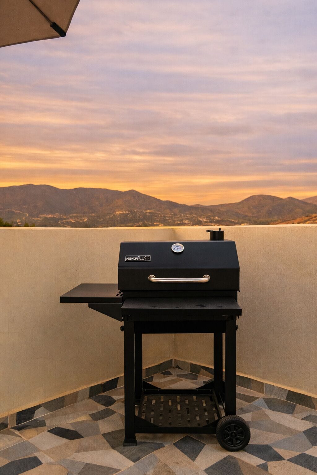 A black grill with a handle and a logo, placed on a tiled patio with a view of mountains and a lake in the background.