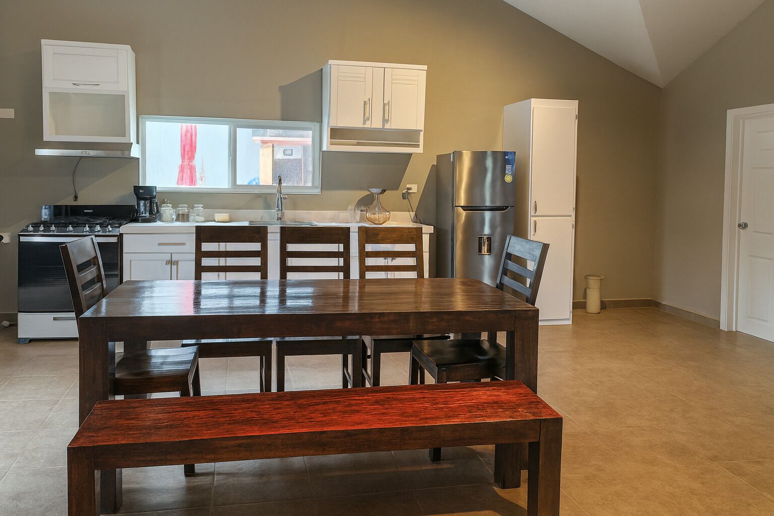 A modern kitchen with a dining area, featuring a stove, refrigerator, cabinets, and a large window with curtains.