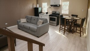 Een modern appartement met een grijze bank, een eettafel met stoelen, een keuken met een fornuis en magnetron, en een houten trap.